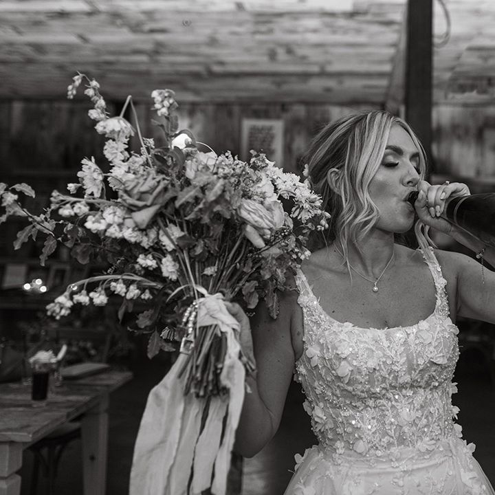 Bride in floral applique and embellished wedding dress holding her oversized wedding bouquet drinking champagne from the bottle 