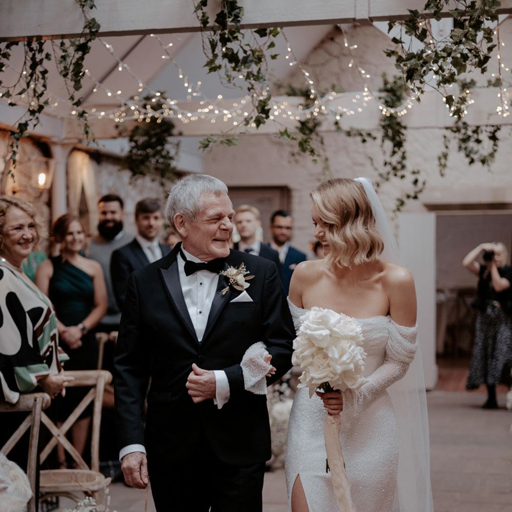 Father of the bride in black tuxedo walks the bride in an off the shoulder sparkly wedding dress down the aisle holding a white rose bouquet 