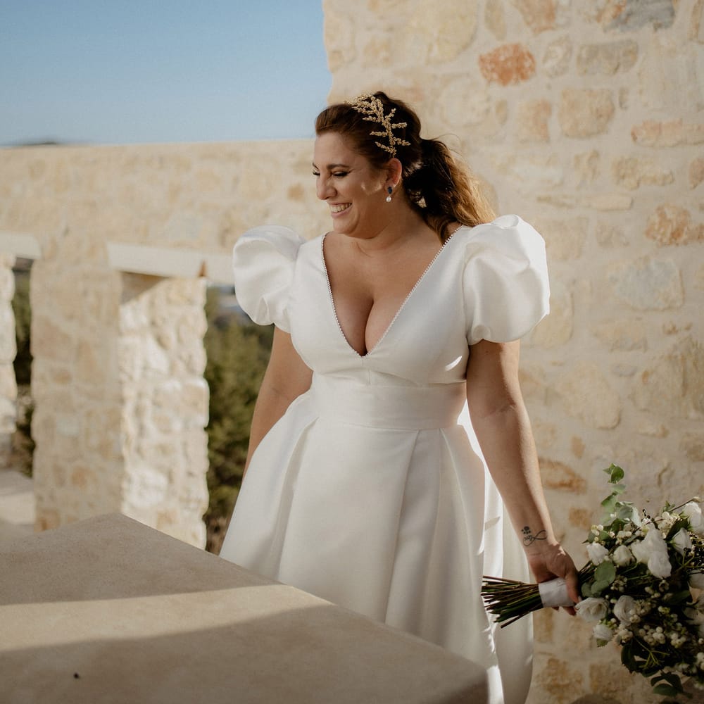 Bride laughing and holding bridal bouqet that features wihte berries, italian ranunculus and eucalyptus whilst wearing a stain puff sleeved wedding dress, peal earrings and gold leaf bridal grown