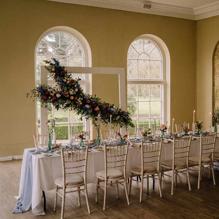Classic wedding tablescape with white wedding tablecloth, cornflower blue wedding table runner, bright blue glass bottles with dried flowers, pastel tapered candles and spring floral arrangements with suspended spring floral decor 