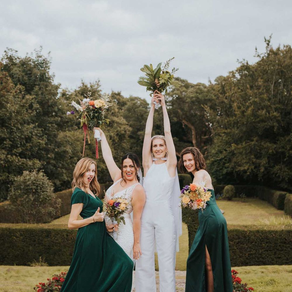 Bride in sleeveless bridal jumpsuit with cape standing with bridesmaids in bottle green velvet bridesmaid dresses 