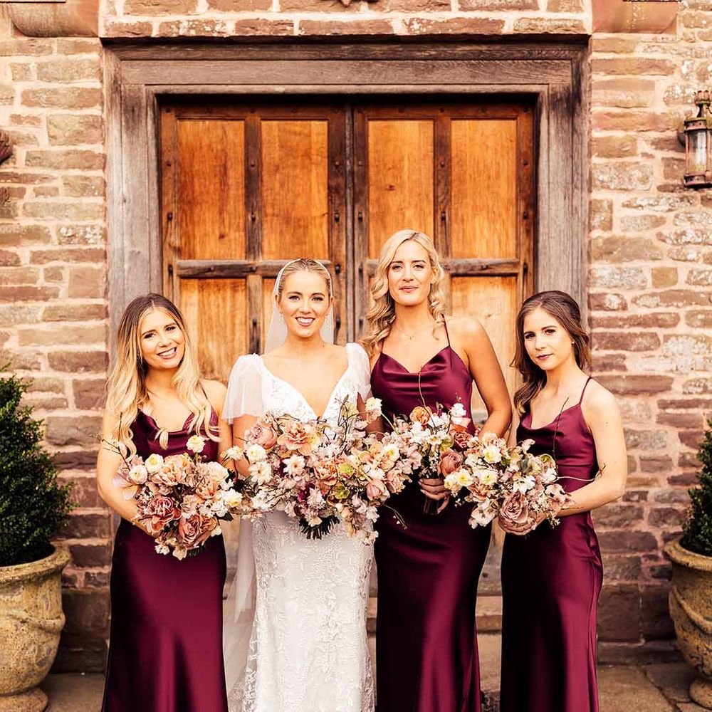 Bride in satin v neck short sleeve wedding dress standing with bridesmaids in strappy burgundy bridesmaid dresses 