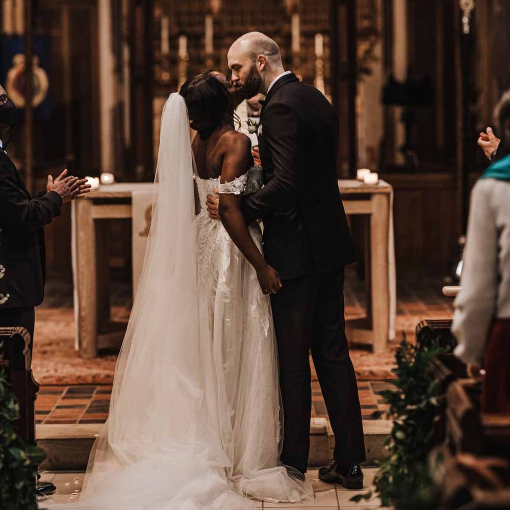 Bride in sweetheart neckline lace wedding dress with detachable off the shoulder sleeves standing with groom in classic black three piece grooms suit with bow tie and white rose and foliage boutonniere in church 