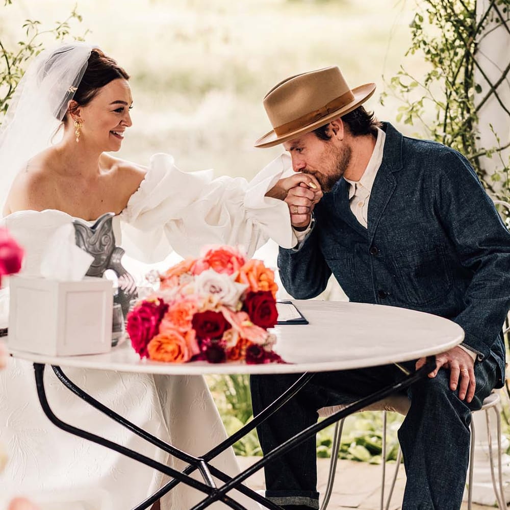 The bride in a puff sleeve gown signs the wedding register with the groom in a double denim suit and hat 
