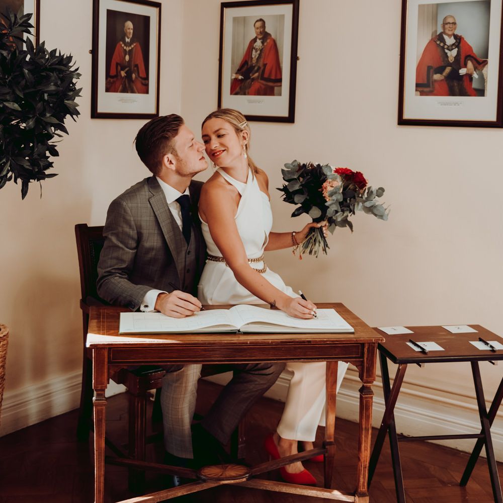 Bride in Massimo Dutti jumpsuit sitting on her grooms lap signing the register 