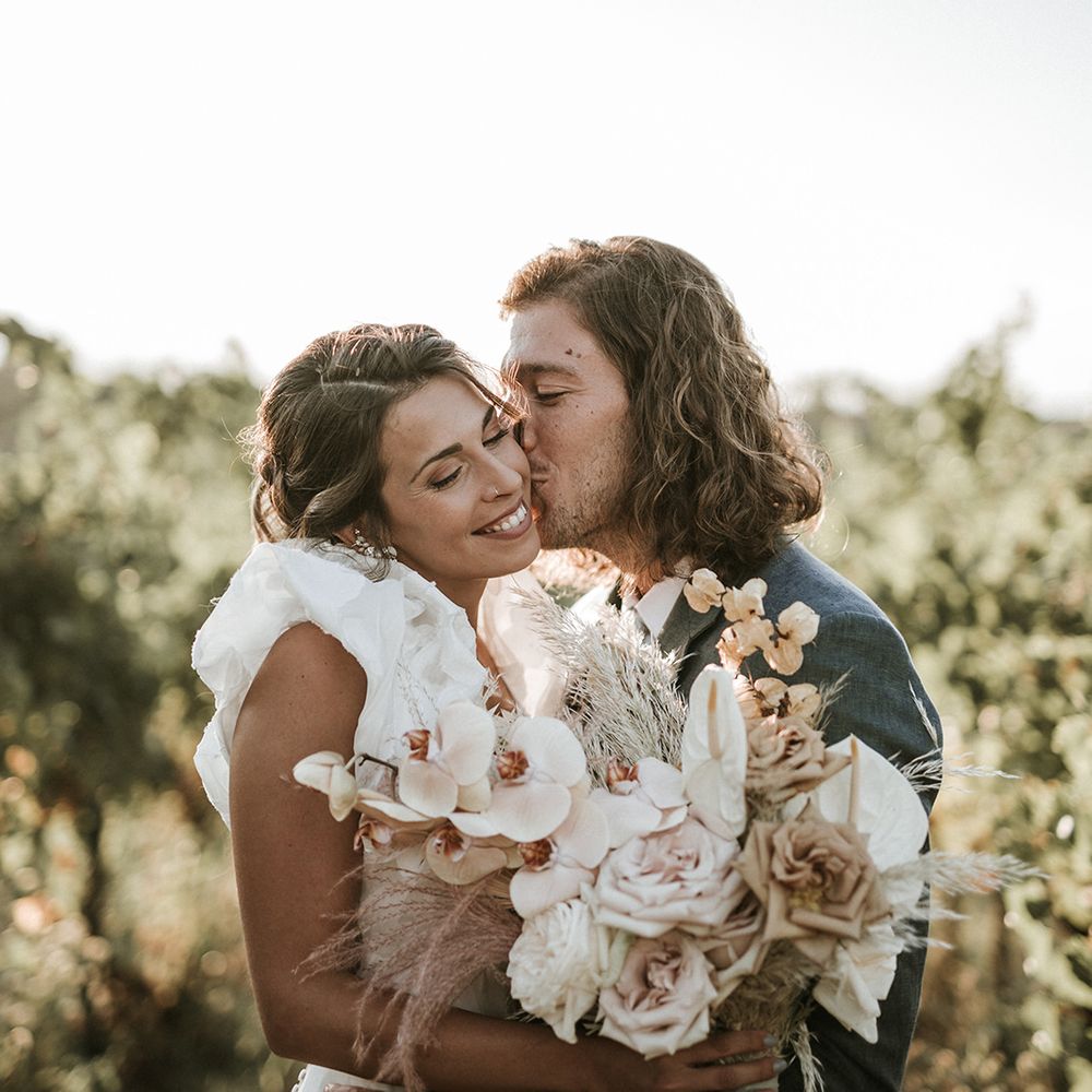 Groom with shoulder length curly hair in a navy blue suit kissing his brides cheek in a ruffle sleeve wedding dress holding an orchid wedding bouquet 