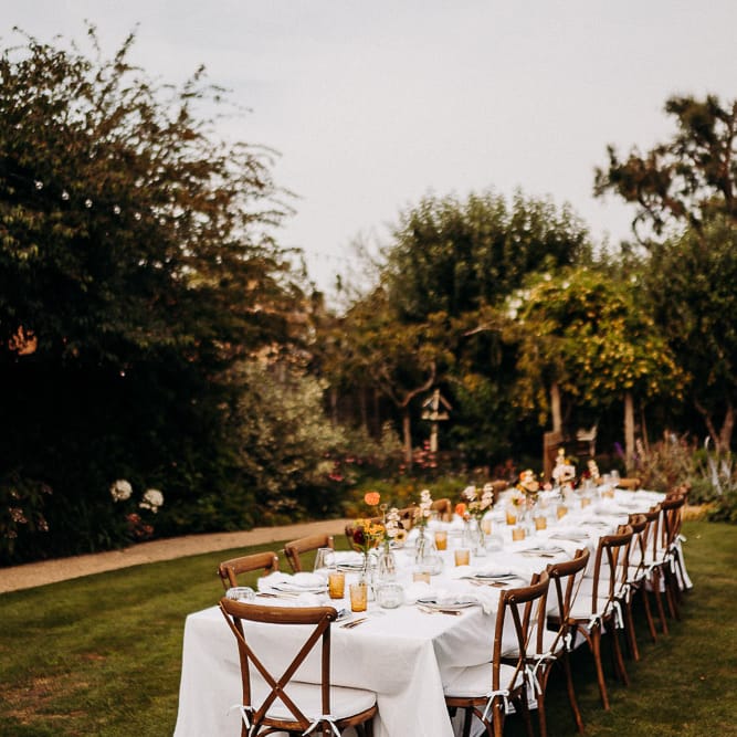 Intimate garden party wedding reception with wooden table and chairs, white table cloth and yellow glassware. 