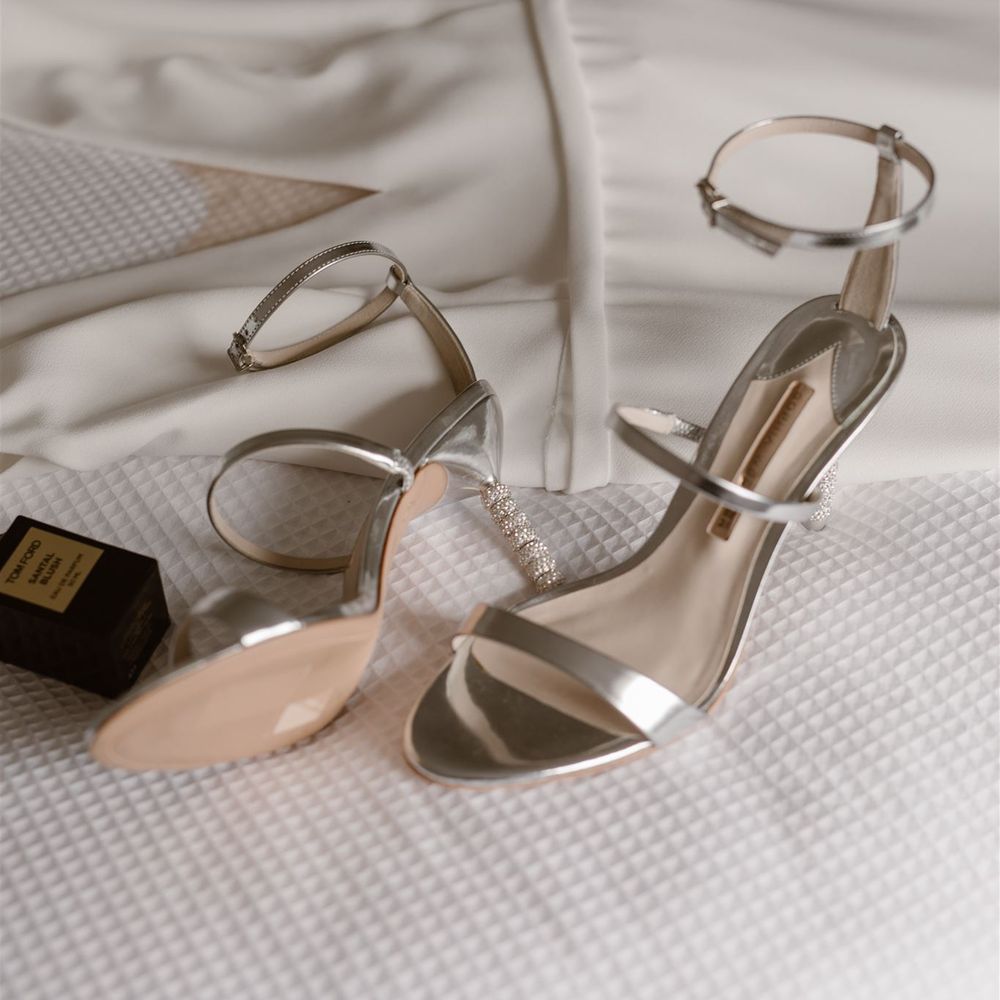 Silver Sophia Webster heeled sandals with three straps and studded heel on white bedspread with black bottle of Tom Ford Eau de Parfum