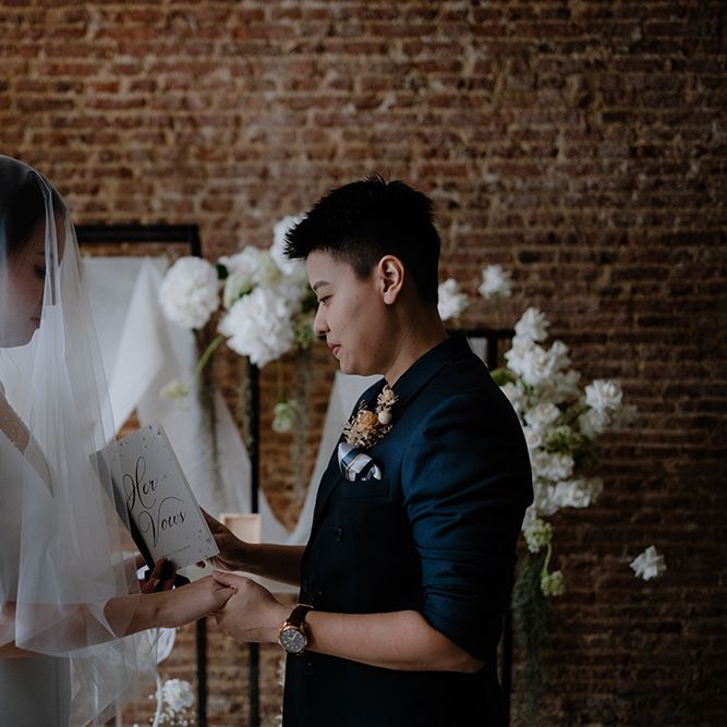 Asian bride in a navy suit jacket reading her vows from a vow booklet 