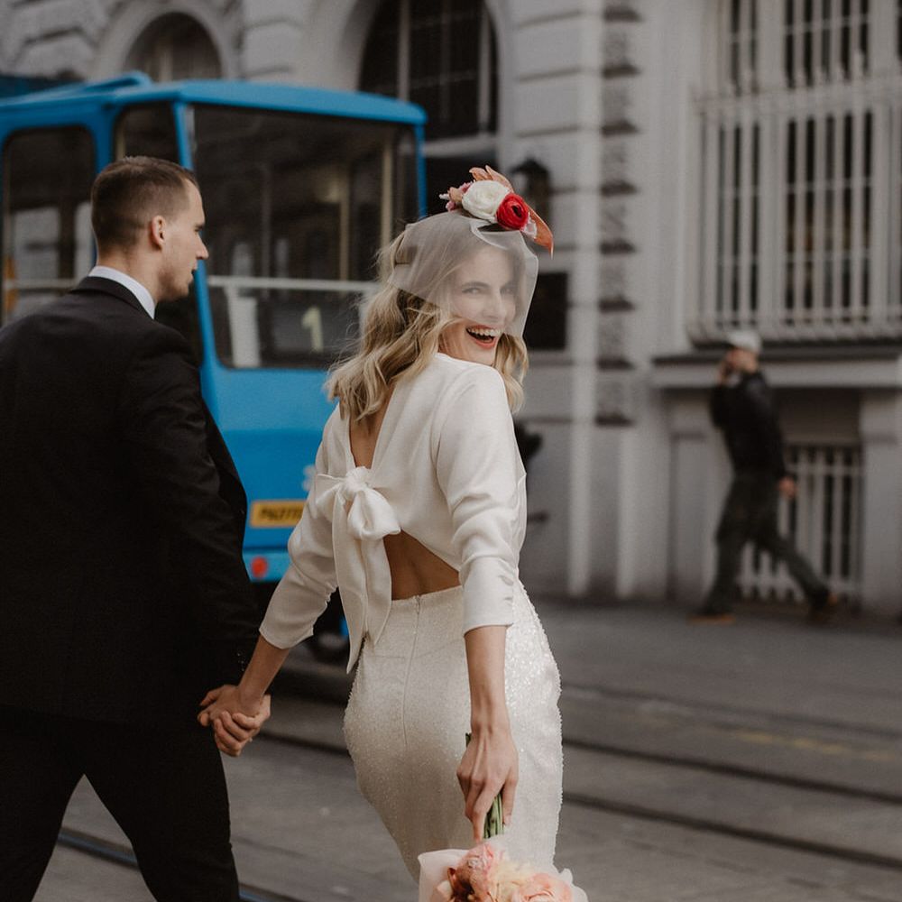 Bride and groom walking through the city, the bride is wearing an open back wedding dress with a bow and sequin pencil skirt by Gray Bridal
