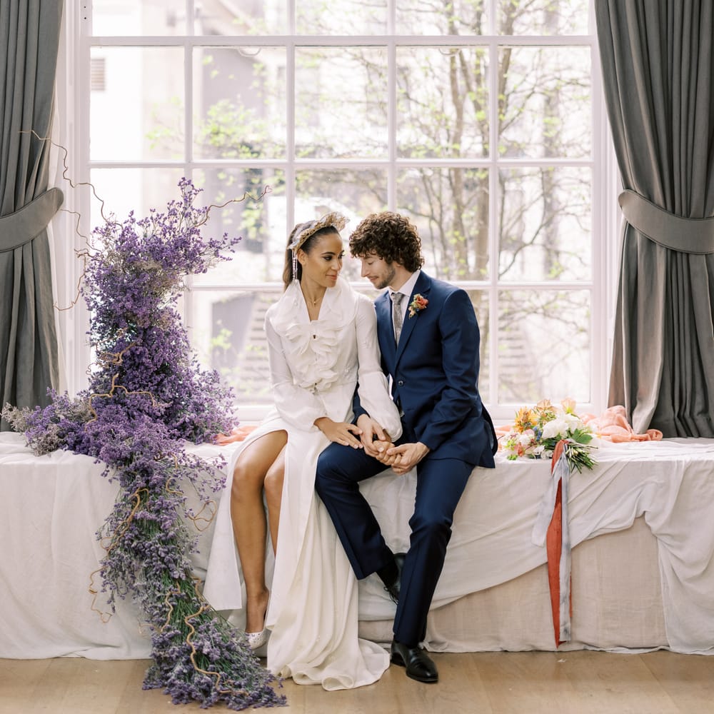 Bride & groom sit on table next to twisted lavender whilst holding hands and looking lovingly at one another