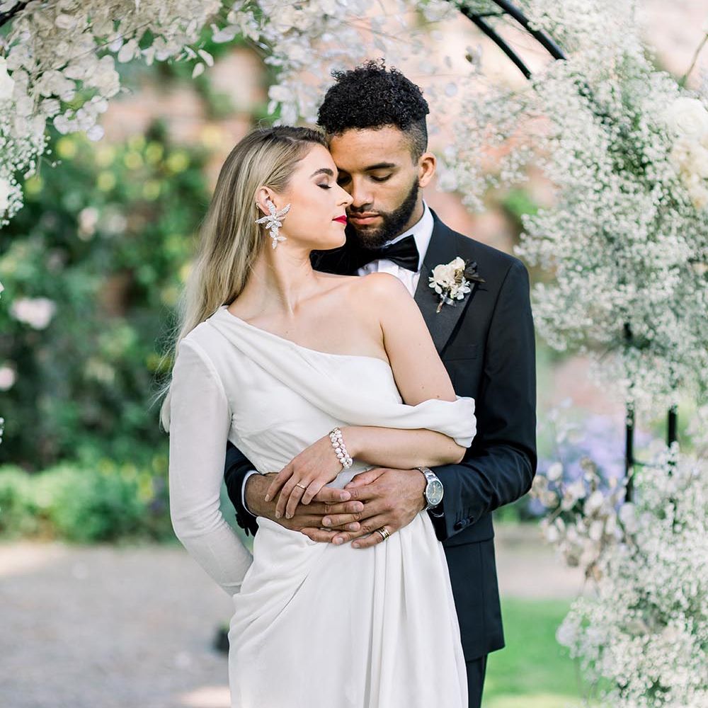 Groom in a black tuxedo embracing his bride in a one shoulder wedding dress with long sleeve and statement earring 