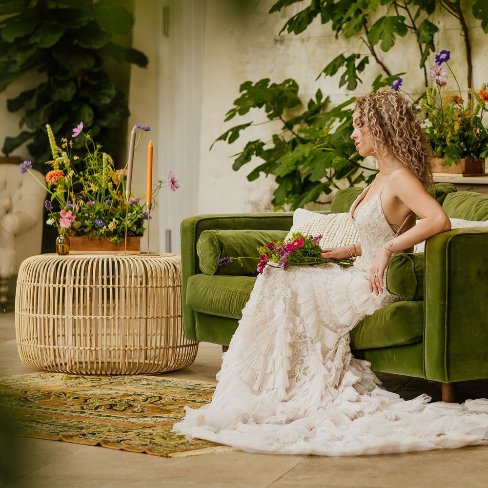 Blonde curly haired bride sitting on a green velvet sofa in a embellished wedding dress surrounded by colourful wildflower decor 