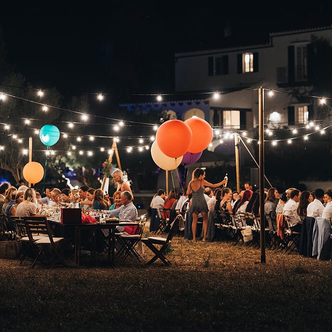 Wedding reception during the nighttime in Menorca with fairy lights strung above wooden tables and colourful balloons