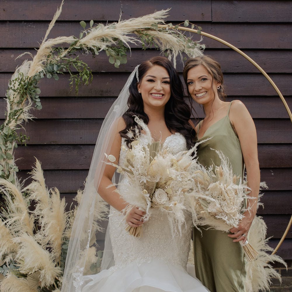 Bride in a fishtail wedding dress holding a dried flower wedding bouquet standing in front of a pampas grass hoop with her bridesmaid in a green satin dress