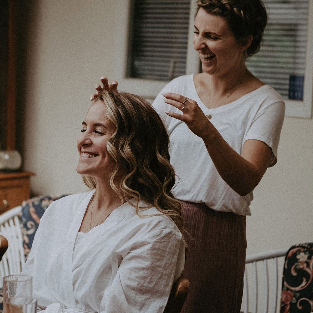 Bride with curled hair smiles as hairdresser arranges fringe sat in kitchen before Isle of Wight wedding with macrame wedding decor