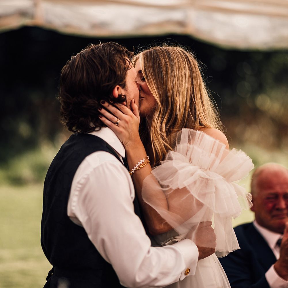 Groom in white shirt and blue waistcoat kisses bride in white Halfpenny London Mayfair dress during garden marquee wedding reception in Cornwall