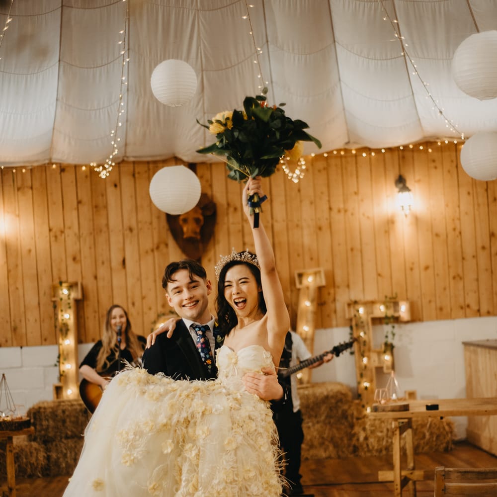 Groom in dark suit and floral tie carries bride in strapless feathered wedding dress and headpiece as she holds up bouquet during rustic wedding breakfast with fairy lights