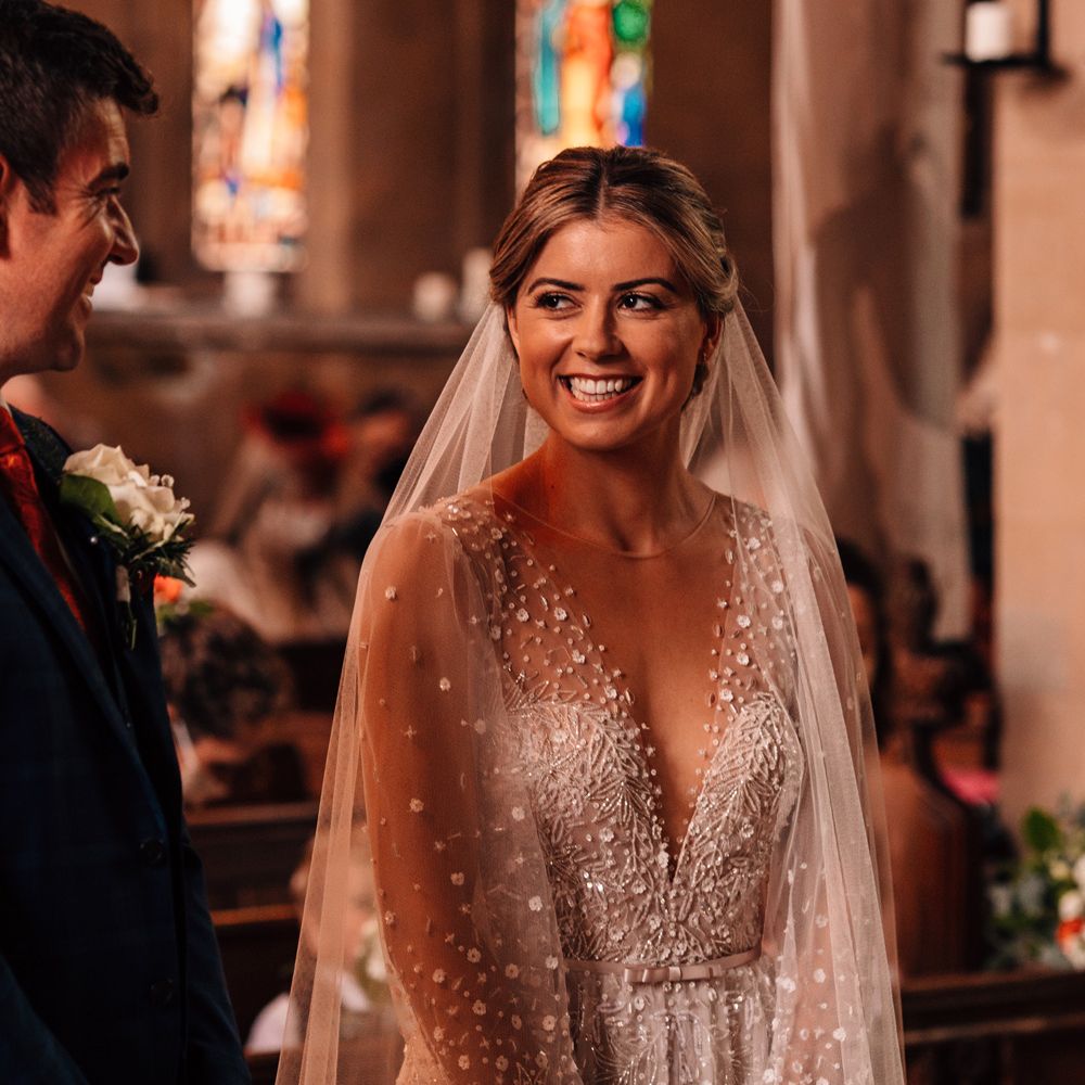 Bride in plunging v neck Julie Vino wedding dress and veil smiles at groom in checked blue suit and white buttonhole curing church wedding ceremony