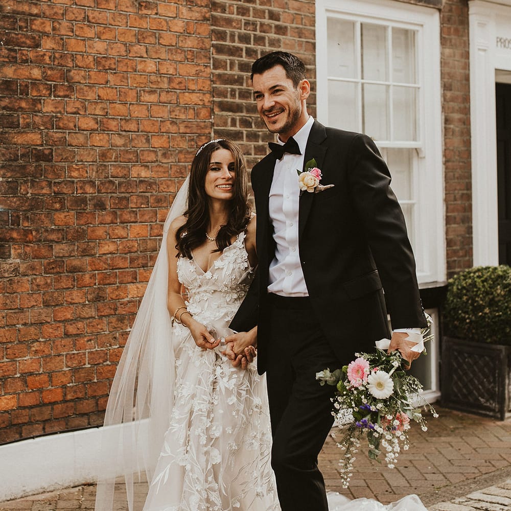 Bride & groom hold hands as they walk together and groom holds floral bouquet