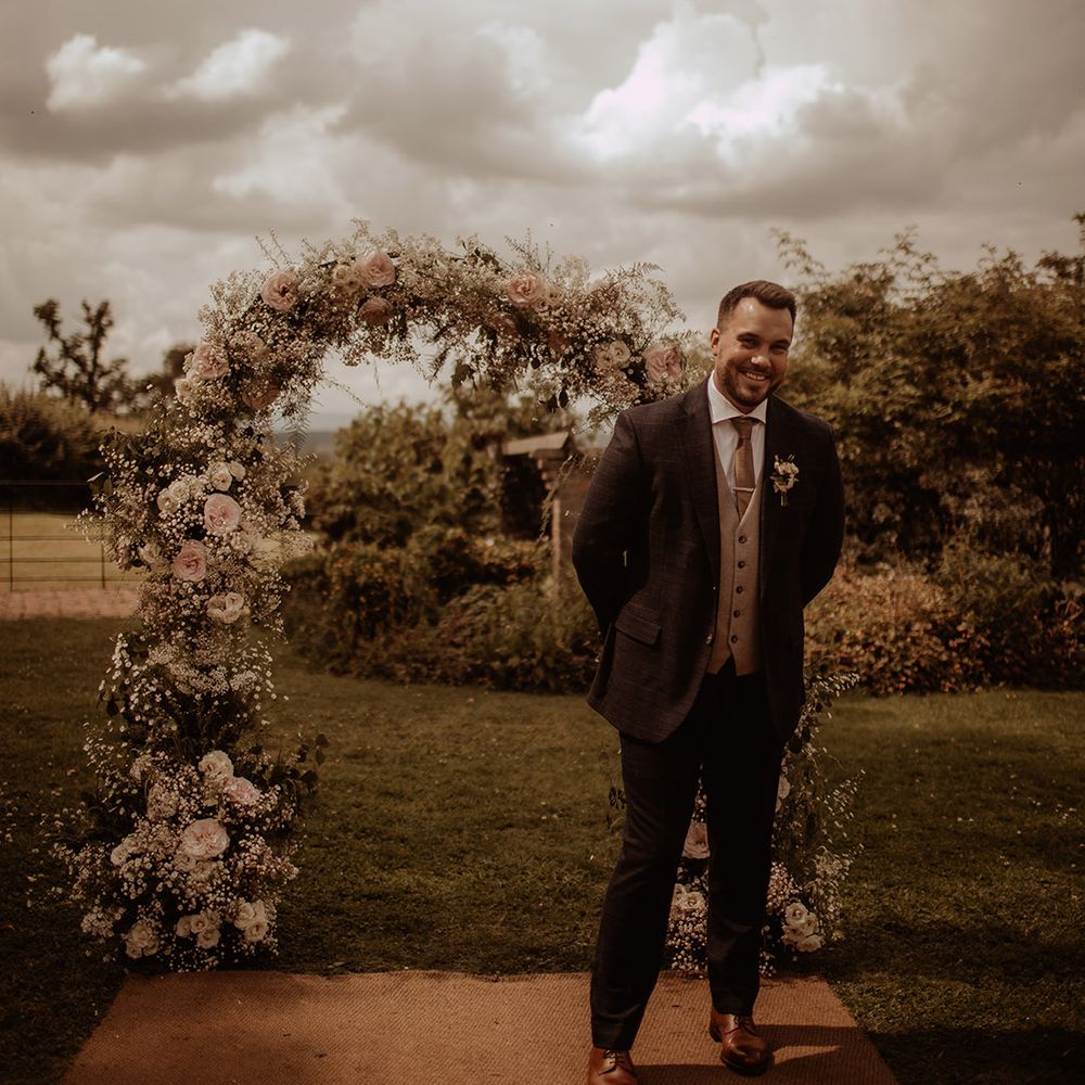 Groom in a three piece suit waiting at the altar with a pink rose and white gypsophila flower arch 