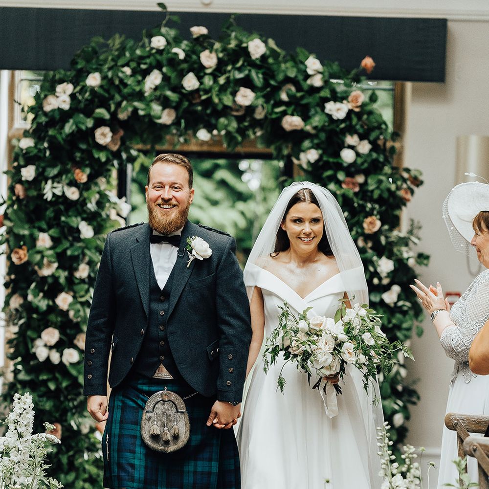 Bride in an off the shoulder Suzanne Neville wedding dress and groom in a tartan kilt standing in front of a floral arch