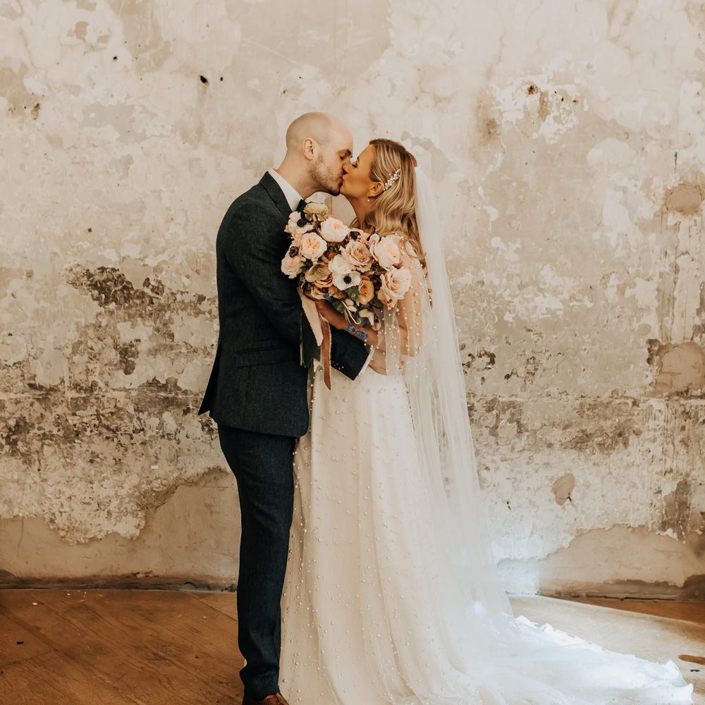 Bride in white wedding dress with pearl mesh overlay holding mixed rose and anemone bridal bouquet kisses groom at industrial wedding venue