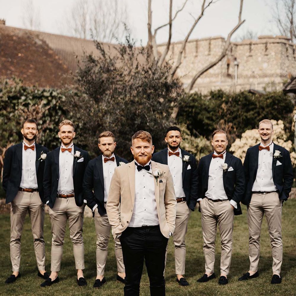 Groom in beige suit jacket and black bow tie and trousers and groomsman in light coloured trousers and orange bow ties