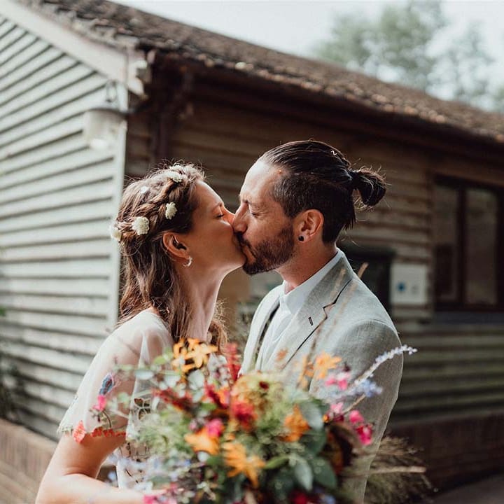 Bride and groom share a kiss outside their wedding venue with bride holding colourful wedding bouquet
