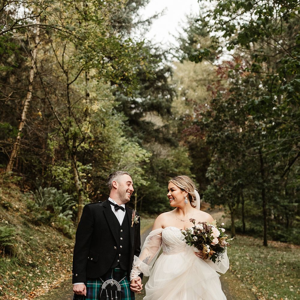 Bride and groom walk a path together holding hands with bride in custom wedding dress and groom in black tie and traditional kilt 