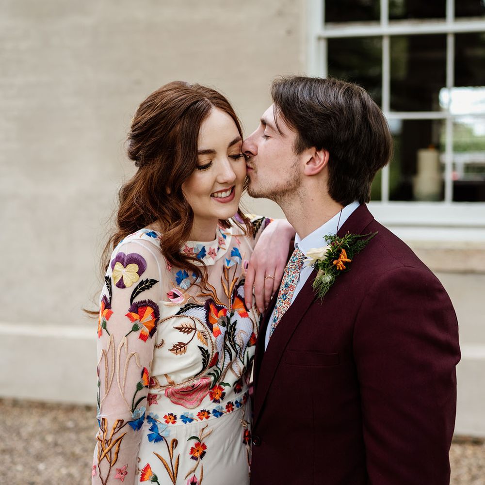 Groom kisses the bride on the cheek as she leans against him wearing bright flower wedding dress 