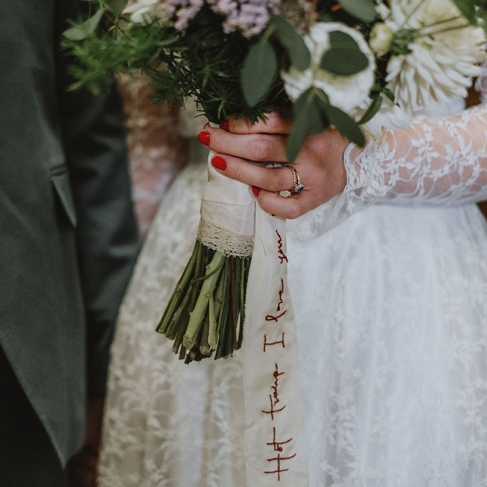 Bride holds sustainable floral bouquet tied with white silk ribbon with personalised stitching in red
