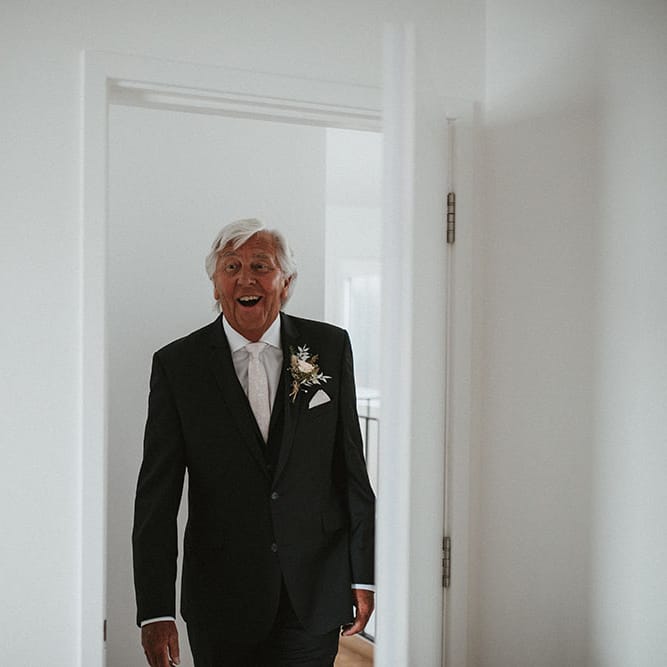 Bride's father wears three piece black suit and floral buttonhole sees his daughter for the first time 
