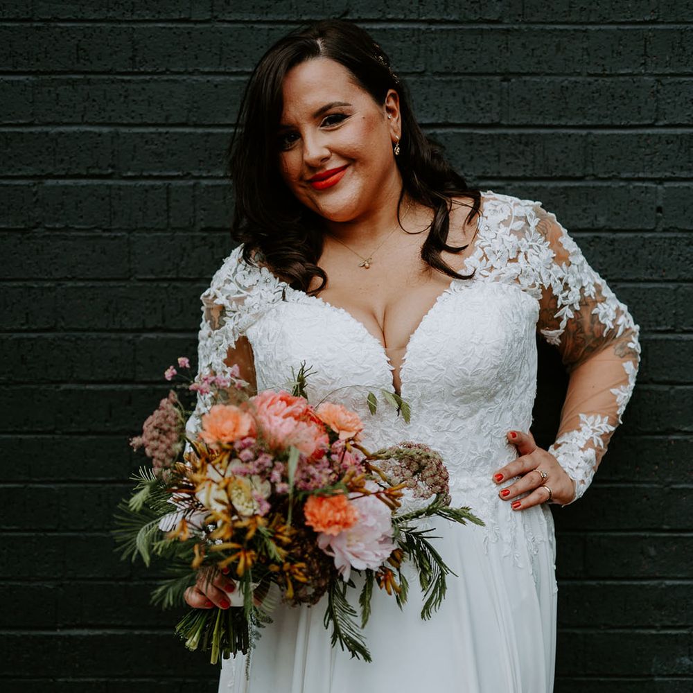 Bride wears lace embellished wedding dress with long sleeves and holds brightly coloured floral bouquet 