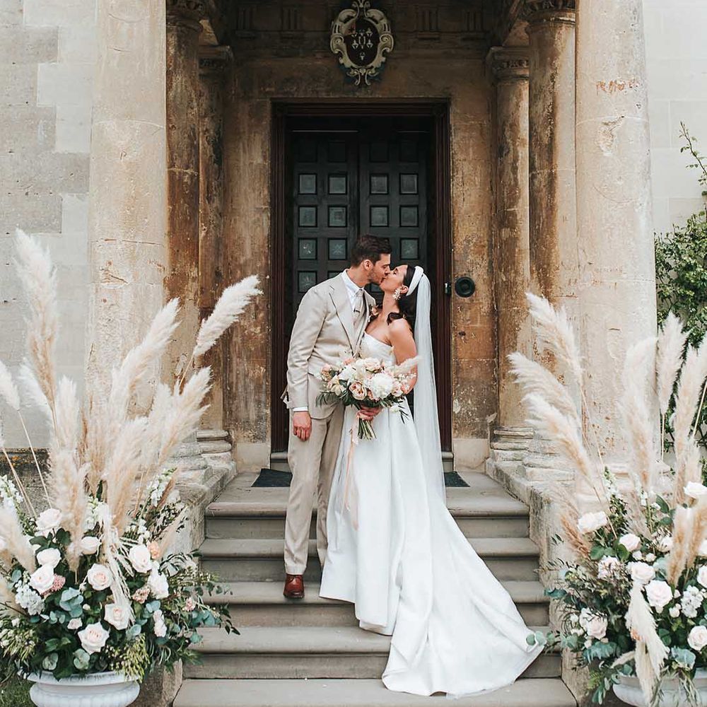 Bride in off shoulder wedding dress with long veil and grey suit embracing on the steps of Elmore Court with pampas grass floral arrangements 
