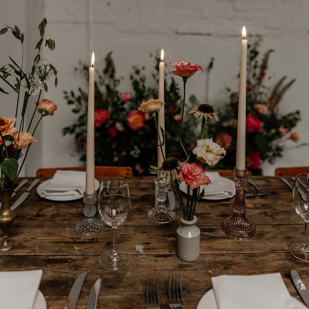 Three tapered ivory candles in clear and rose gold candle holders on neutral minimalistic wedding tablescape with pops of colourful flowers