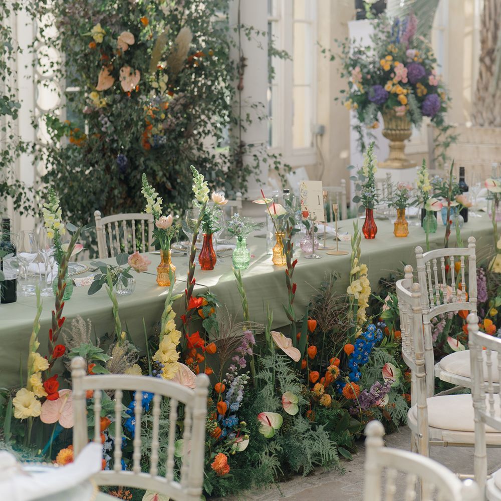 Brightly coloured floral arrangements with green foliage in front of head table with pastel green tablecloth and floral installation to back