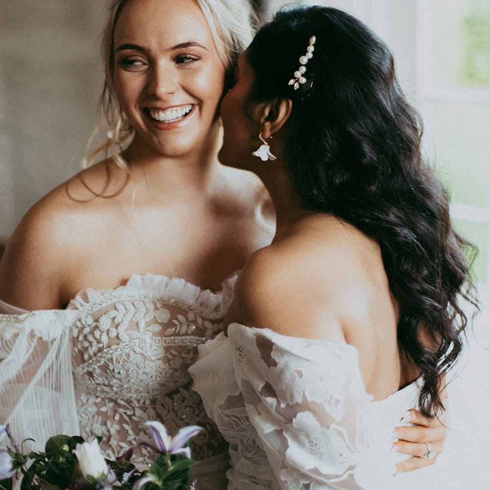 Brides in complimenting lace detailed wedding dresses and pearl hair accessories smiling