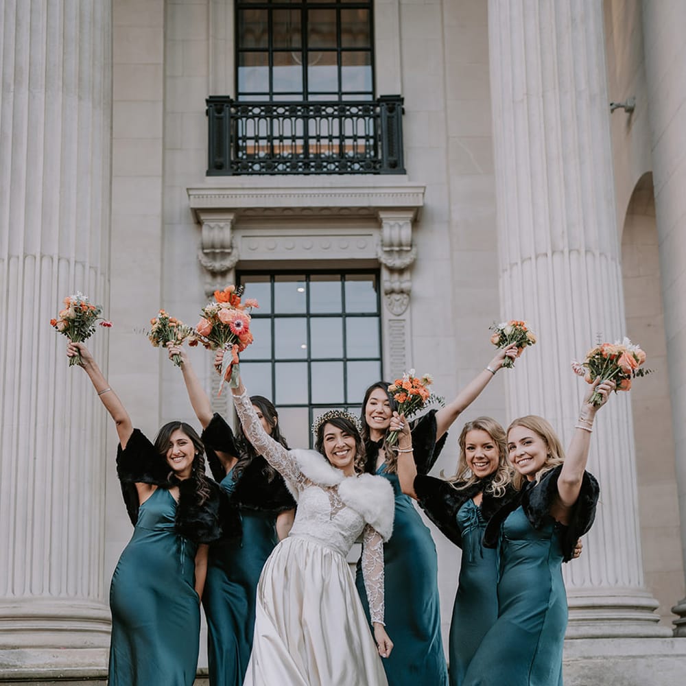 Bride in taffeta material princess wedding dress with long lace sleeves  and cropped faux fur bridal cover up holding up bridal bouquet with bridesmaids in deep green satin bridesmaid dresses with faux black coverups and black open toe heels 
