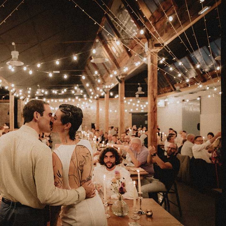 Lisa & Gabriel kiss in front of wedding guests during reception in industrial setting with festoon lighting 