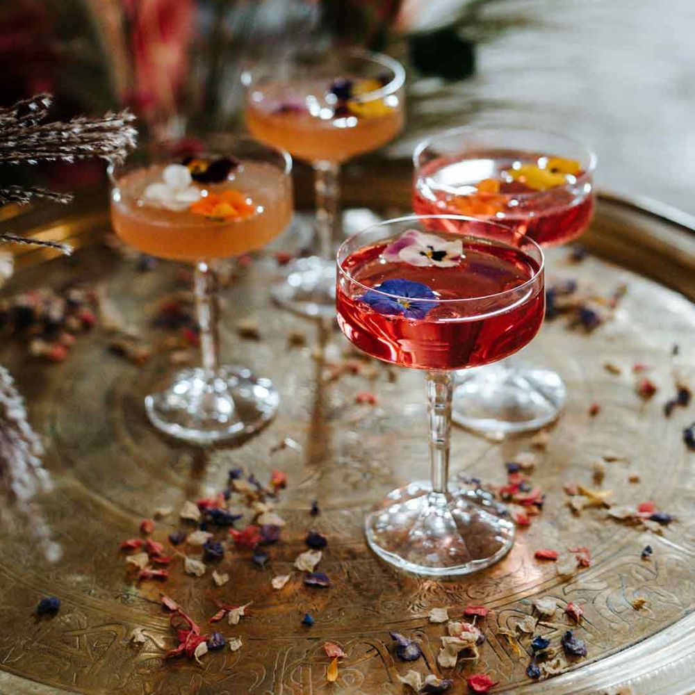 Four colourful wedding cocktails with edible flower toppers in cocktail glasses on wooden slate