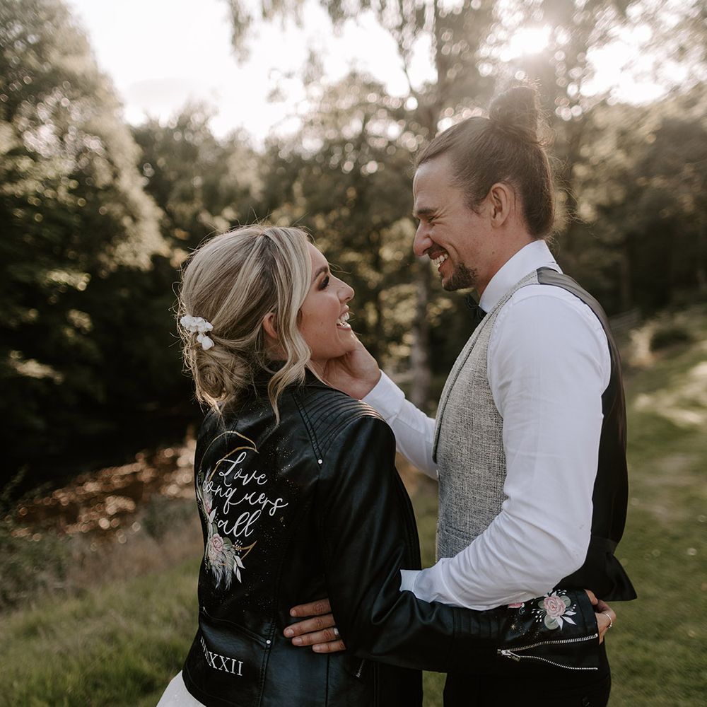 The bride with her blonde hair in an updo in a personalised leather jacket embracing the groom in light grey waistcoat 