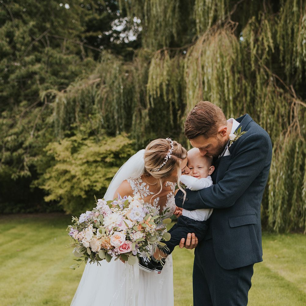 The bride in a Dando London wedding dress kissing the baby boy held by the groom in a navy suit 