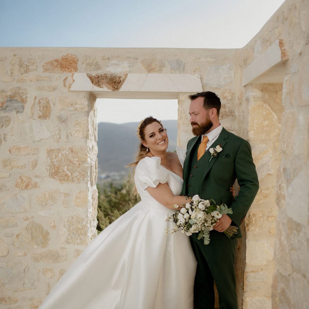 Bride in white satin princess dress featuring puff sleeves poses with groom in dark green wedding suit at private villa wedding in Greece