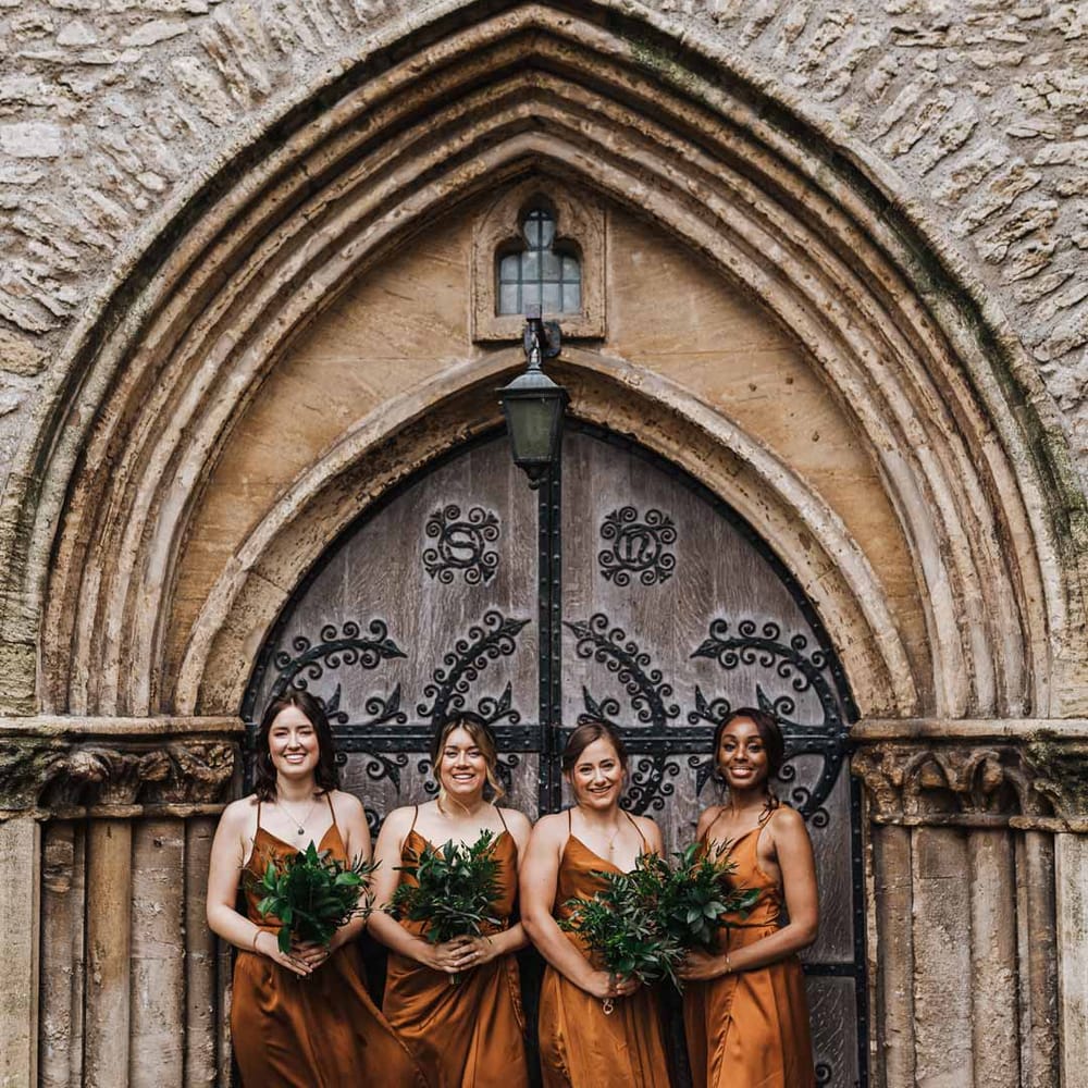 Bridesmaids standing outside of the church wearing strappy sleeveless front slit burnt orange bridesmaid dresses holding foliage bouquets 
