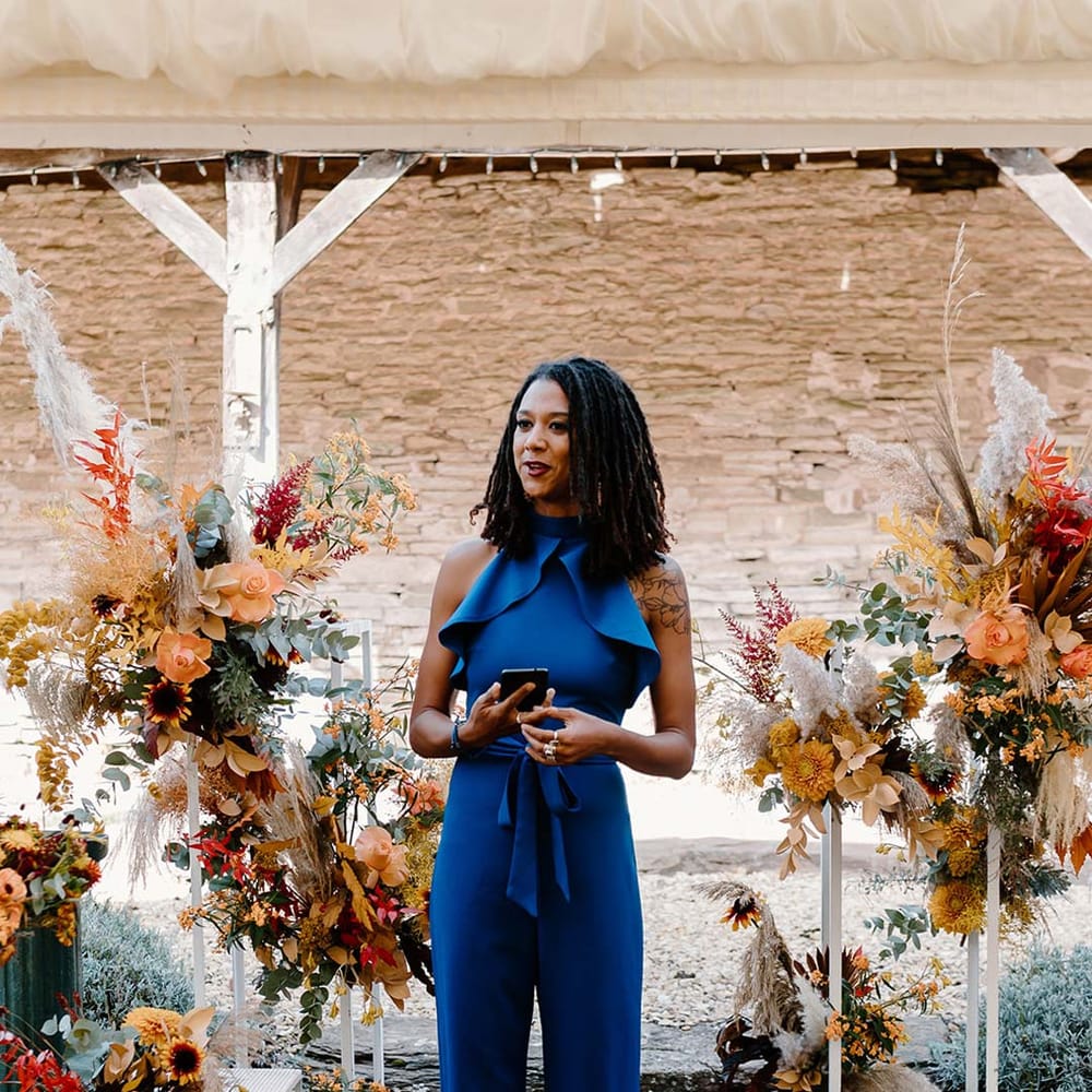 Wedding guest leading the wedding ceremony wearing cobalt blue jumpsuit standing in front of wedding alter flower arrangements with sunflowers, garden roses, pampas grass, eucalyptus, yellow tall kangaroo paws, orange osmanthus flowers, foliage and dried flowers 