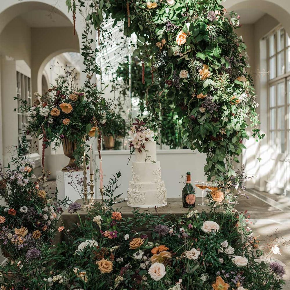 Four tier white wedding cake with intricate icing details and dried flower wedding cake topper on wooden table with twisted tapered candles in neutral shades in gold candlesticks surrounded by wildflower and foliage wedding flower arrangements