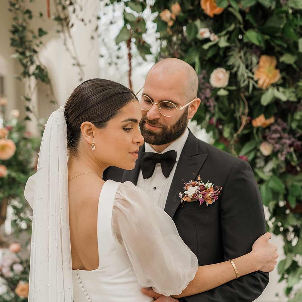 Bride in short sheer puff sleeve wedding dress with square neck and wrist loop with sheer pearl embellished bridal veil with button detailing dancing with groom in classic black tuxedo with bowtie and wildflower boutonniere