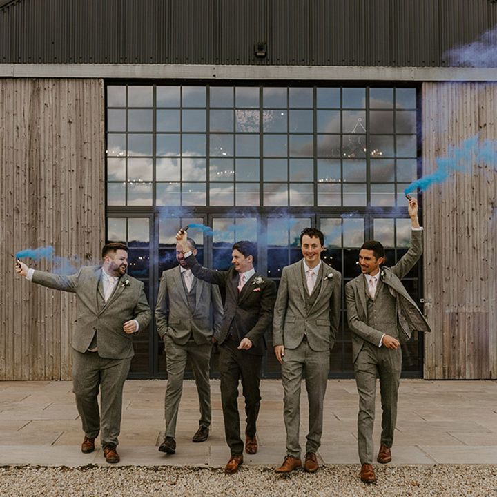 Groomsmen in light grey suits walking with groom waving blue smoke flare 