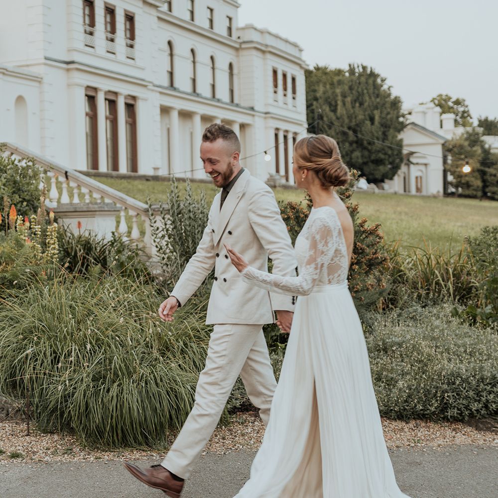 bride in a Catherine Dean floaty skirt wedding dress with front slit and lace long sleeves holding hands with her groom in a beige suit 